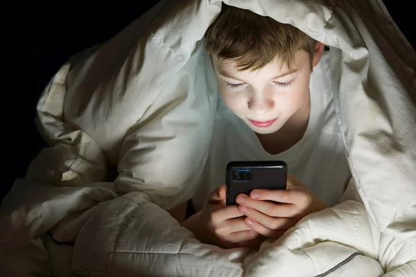 Menino debaixo do cobertor à noite em sua cama se comunica na Internet. Vício em gadgets infantis e insônia. vício em redes sociais — Fotografia de Stock