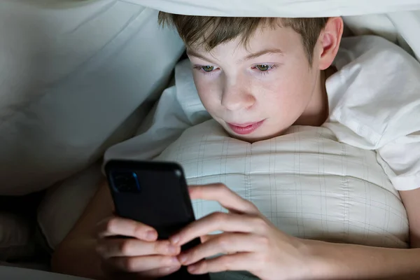 Menino debaixo do cobertor à noite em sua cama se comunica na Internet. Vício em gadgets infantis e insônia. vício em redes sociais — Fotografia de Stock