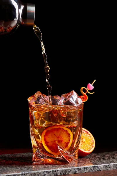 Alcoholcocktail van shaker naar gless met sinaasappelschil-ijs op stenen bureau. — Stockfoto