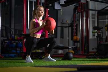 Genç, güzel bir kadın, spor salonunda sağlık topu pozisyonunda antrenman yapıyor..