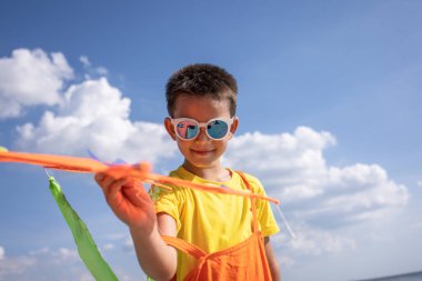 Renkli bir kıyafet içinde güneş gözlüğü takan bir çocuk açık bir günde uçurtmayı yüzdürür..