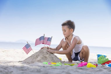Kumsalda oyuncak oynayan sevimli bir çocuk. Yanında Amerikan bayrağı olan bir kum yığını..
