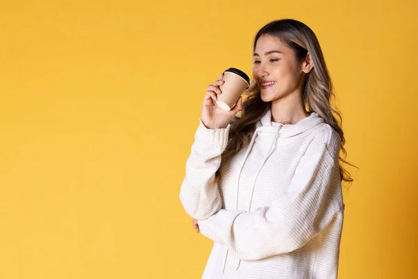 Portrait of Asia woman blond teenage girl with wavy hair looking charming holding paper disposable cup for coffee on over yellow background.