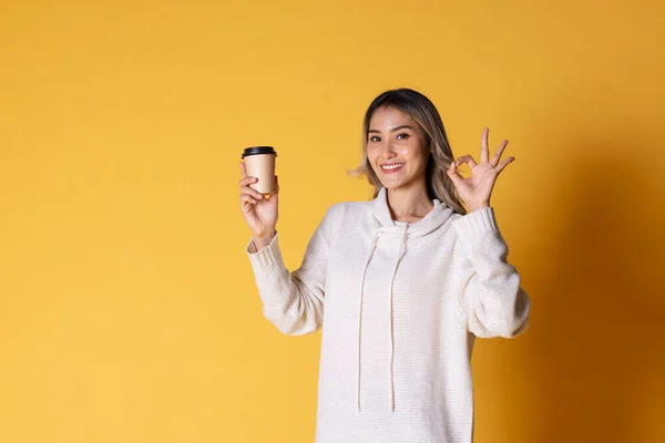 Portrait of Asia woman blond teenage girl with wavy hair looking charming holding paper disposable cup for coffee on over yellow background.