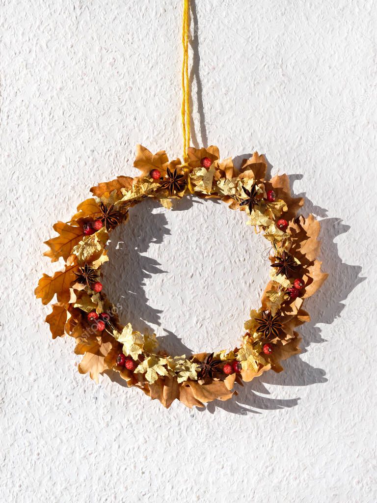 Dried floral wreath made of dry Autumn leaves and berries hang on off white wall with textured wallpaper on yellow cord. Direct sunlight with shadows.
