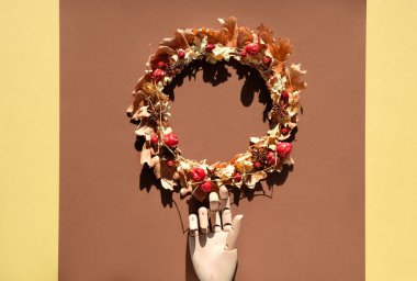 Kurumuş güz yapraklarından yapılmış kurumuş çiçek çelengi ve kahverengi ve sarı katmanlı kağıt arka planda böğürtlenler. Manken tahta el çelengi tutar. Gölgeli doğrudan güneş ışığı.