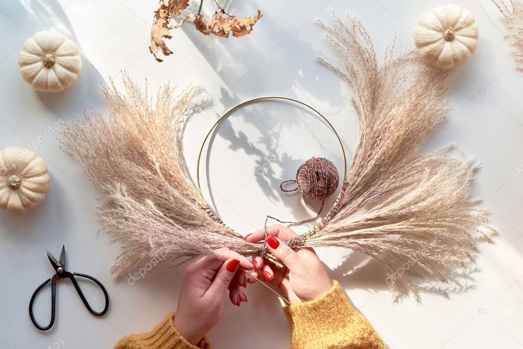 Hands making dried floral wreath from dry pampas grass and Autumn leaves. Hands in sweater with manicured nails tie decorations to metal frame. Flat lay on off white table, sunlight with long shadows.