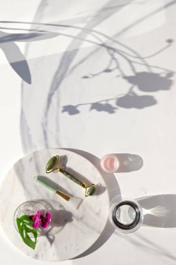 El masajı, yeşil taş silindirli lenf drenajı. Mermer tabakta nemlendirici krem. Canavar yapraklı egzotik bir arka plan, canlı morumsu orkide çiçekleri. Güneş ışığı, uzun gölgeler, üst manzara.