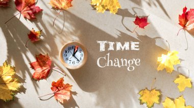 Sonbaharda zaman değişiyor. Tahta alarm saati, bir saat işaretli ve geri dönüşümlü karton üzerinde kuru yapraklar var. Tahtada metin zamanı değişimi. Üst manzara, düz uzanma, uzun gölgeli güneş ışığı..