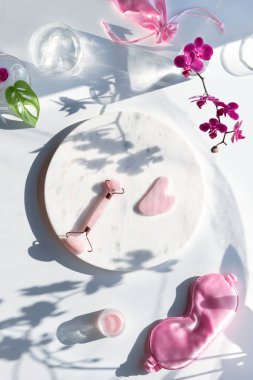 Pembe kuvars yüz silindiri, mermer tabakta nemlendirici krem. Canavar yaprakları ve morumsu orkide çiçekleriyle egzotik düz yatıyordu. Güneş ışığı, uzun gölgeler. Yüz masajı.