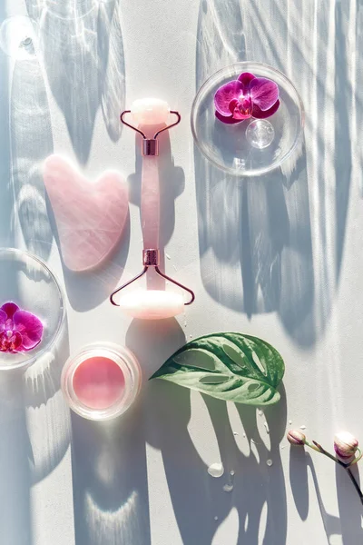 Pink quartz face roller and guasha stone. Face lymph drainage background with exotic monstera leaves, orchid flowers, petri dishes, glass balls. Sunlight, long shadows. Flat lay off white background.