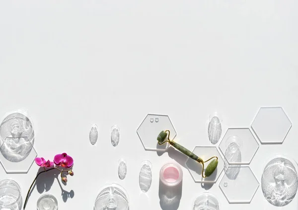 Jade stone face roller with fuchsia orchid flowers and glass baubles. Pink moisturizer cream in jar. Sunshine, long shadows. Flat lay on off white background with copy-space. Face lymph drainage.
