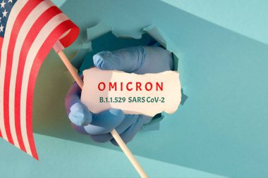 Amerika Birleşik Devletleri 'ndeki Omicron. Yeni bir korona virüsü türü. Yırtık kağıt deliğinden elde tutulan eldiven ABD, Amerikan bayrağı ve yeni Coronavirus varyantının adı ile bir kağıt parçası içeriyor..