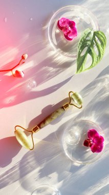 Canavarımsı yaprağı, orkide çiçekleri ve cam süslemeleri olan yeşim taştan bir silindir. Kavanozda pembe nemlendirici krem. Güneş ışığı, uzun gölgeler. Güzellik tedavisi düzlüğü, kopyalama alanı olan beyaz arkaplan üzerinde yatıyordu.