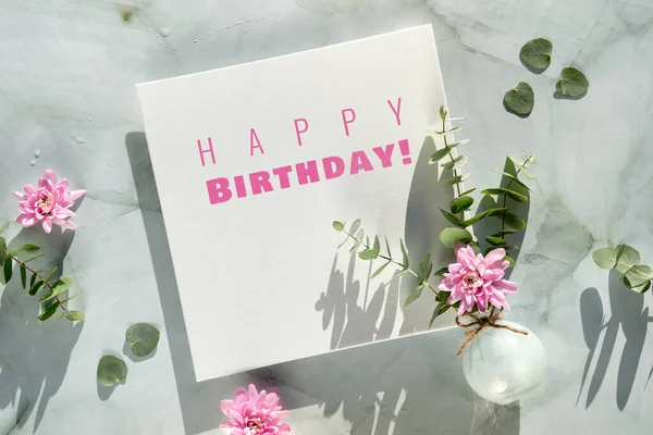 Happy birthday text on square canvas and winter, early spring decorations. Pink chrysantemum flowers, eucalyptus in glass vase on mint green stone. Greeting card design in trendy pastel colors.