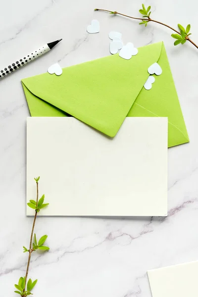 緑の色合いの春の背景 空白のグリーティングカード コピースペースと紙の封筒 新鮮な葉を持つ春の小枝 白の背景にハートのコンフェッティ — ストック写真