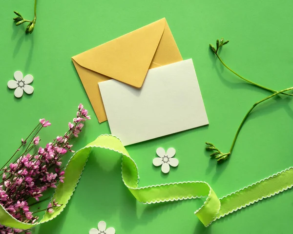 緑と黄色の色合いの春の背景 白紙のカード コピースペースと紙の封筒 緑の紙の背景にピンクの花 デコ木製の花のシルエットと緑のテキスタイルリボン — ストック写真