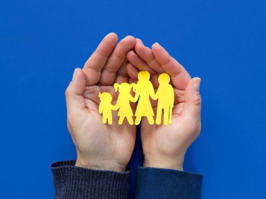 Ukraynalı ailenin sarı kağıt siluetini tutan kadın ve erkek, yardım eden eller. Düz yerde, üst tarafta. Yaratıcı konsept kağıt sanatı mavi ve sarı renklerde. Ukraynalı mültecilere gönüllü yardım.