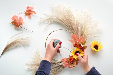 Kuru otlardan ve sonbahar yapraklarından kurumuş çiçek çelengi yapan eller. Manikürlü çivili kazak giymiş eller metal çerçeveye süslemeler bağlıyor. Dümdüz beyaz masanın üzerinde, uzun gölgeli güneş ışığı..