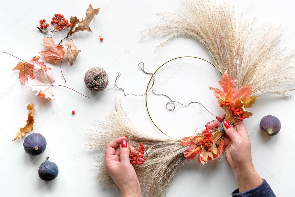 Hands making dried floral wreath from dry pampas grass and Autumn leaves. Hands in sweater with manicured nails tie decorations to metal frame. Flat lay on white table, sunlight with long shadows.