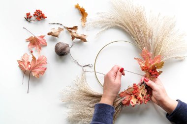 Kuru otlardan ve sonbahar yapraklarından kurumuş çiçek çelengi yapan eller. Manikürlü çivili kazak giymiş eller metal çerçeveye süslemeler bağlıyor. Dümdüz beyaz masanın üzerinde, uzun gölgeli güneş ışığı..
