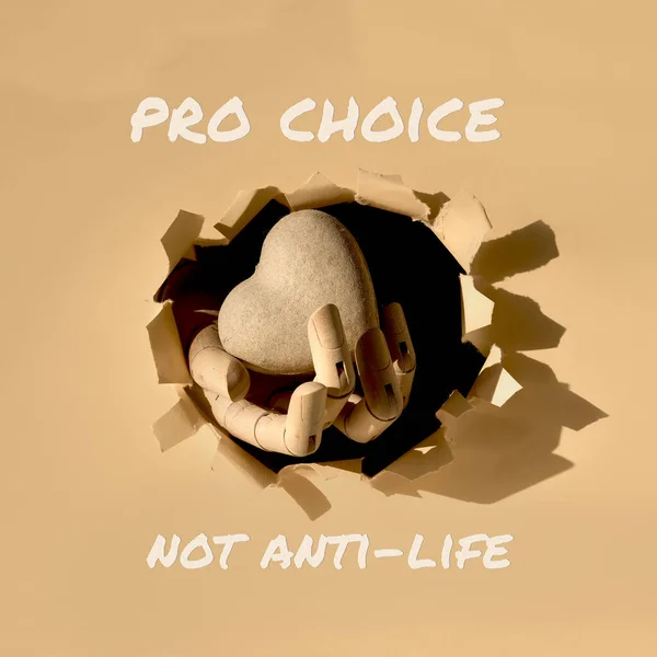 Text Pro Choice not Anti-Life. Cardboard heart in wooden model hand through paper hole in torn beige recycled paper. Roe versus Wade reversal, social dispute concept.