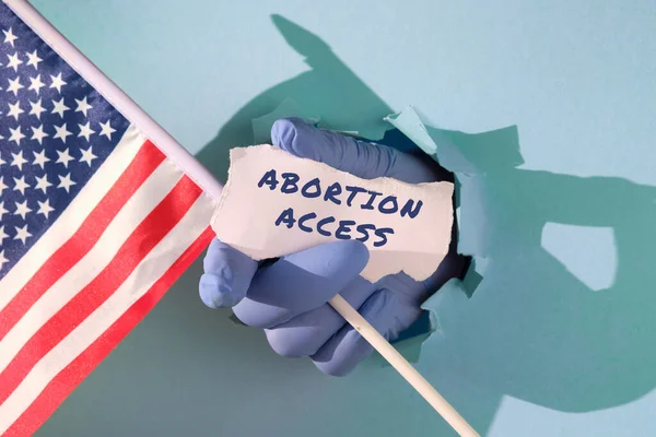 Text Abortion Access on scrap of paper. Reversal of Roe versus Wade decision in United States of America. Medics, doctors hand in glove holding USA american flag and scrap of paper text through paper hole.