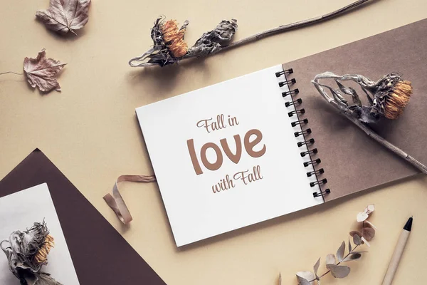Caption motivator text Fall in love with Fall in square album, notebook with spiral binder. Wooden pen, dry sunflower flowers and maple leaves. Soft natural light. Monochromatic beige color hues.