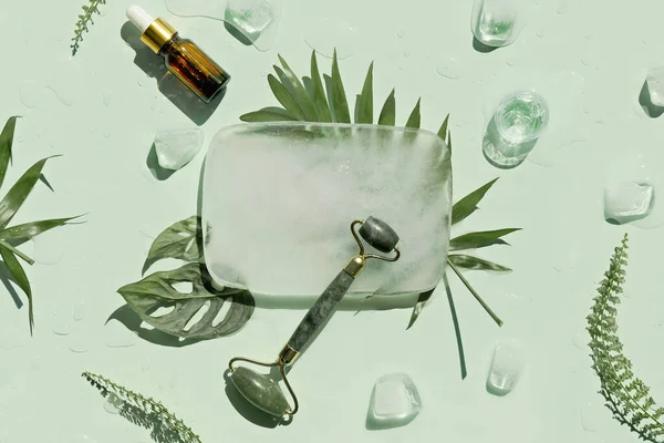 Jade stone face roller, essential oil and handmade cream with ice cubes and exotic palm fern leaves on mint green background. Facial massage concept, handmade cosmetics.