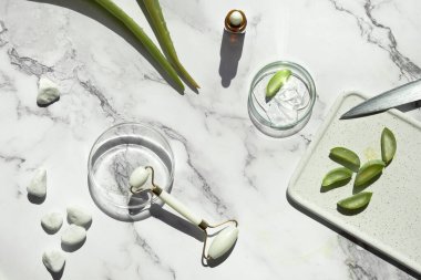 Self made aloe moisturizer and green jade face roller. Exotic fern leaves on off white marble background. Minimal flat lay, top view. Facial massage concept, handmade cosmetics.