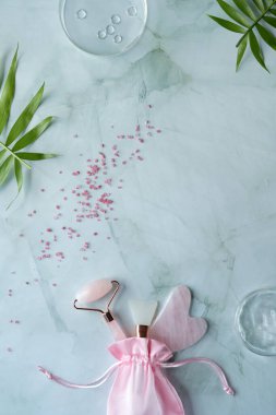 Rose quartz stone facial roller. Facial anti-age massage for natural lifting treatment. Beauty concept, flat lay on mint green with palm leaves. moisturizer in petri dish.