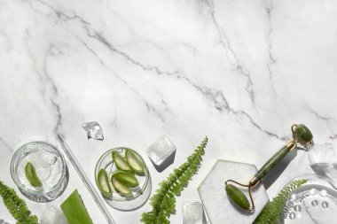 Self made aloe moisturizer and green jade face roller. Exotic fern leaves on off white marble background. Minimal flat lay, top view. Facial massage concept, handmade cosmetics.