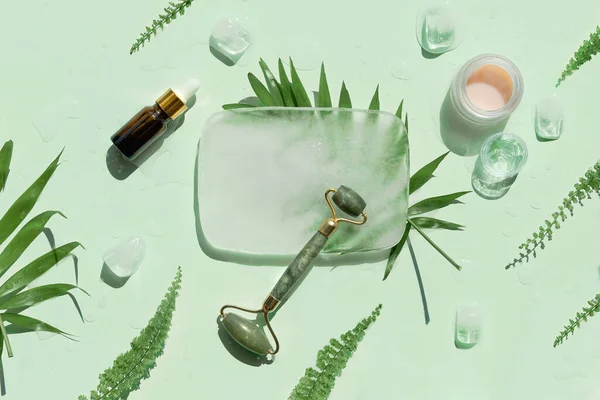 Jade stone face roller, essential oil and handmade cream with ice cubes and exotic palm fern leaves on mint green background. Facial massage concept, handmade cosmetics.