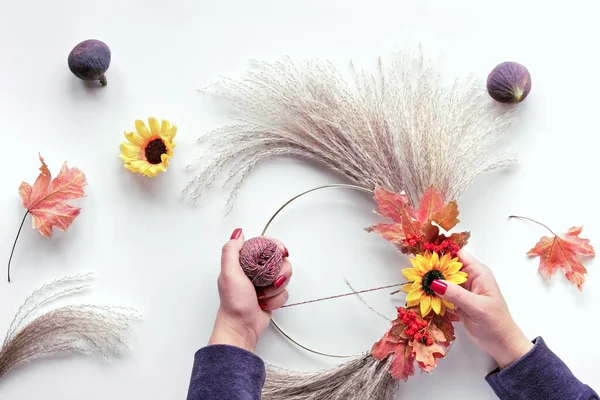 Floral wreath from dry pampas grass and Autumn leaves. Hands in sweater with manicured nails tie decorations to metal frame. Flat lay on white table, sunlight with long shadows.