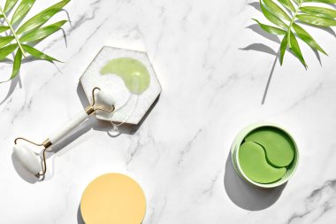 Hydrogel pads and chinese stone roller on off white marble stone background, palm leaf. Direct sunlight, long shadows. Eye patch, skincare beauty treatment at home. Part of skin daily care routine.