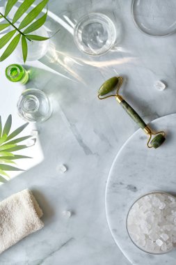 Home made moisturizer and green jade stone face roller on off white marble background. Beauty treatment background, lat lay, top view. Facial massage concept, handmade cosmetics.