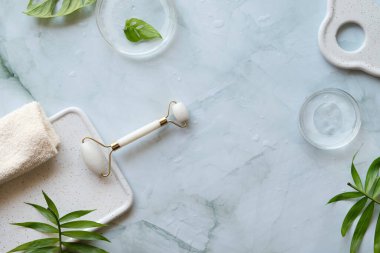 Home made moisturizer and green jade stone face roller on off white marble background. Beauty treatment background, lat lay, top view. Facial massage concept, handmade cosmetics.