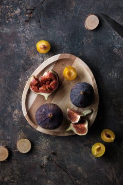 Autumn fruits, black fig and yellow plums on wooden tray. Flat lay, top view on dark textured background with dry poppy plants and hemp cord. Fruits in Fall season.