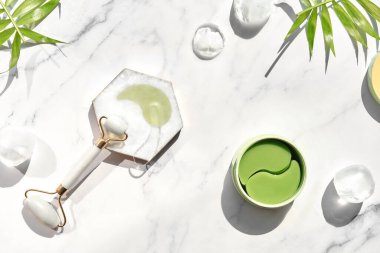 Hydrogel pads and chinese stone roller on off white marble stone background, palm leaf. Direct sunlight, long shadows. Eye patch, skincare beauty treatment at home. Part of skin daily care routine.