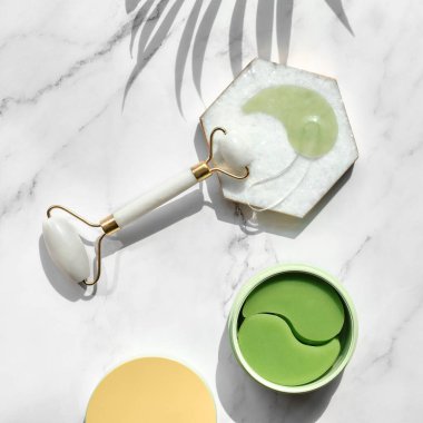 Hydrogel pads and chinese stone roller on off white marble stone background, palm leaf. Direct sunlight, long shadows. Eye patch, skincare beauty treatment at home. Part of skin daily care routine.