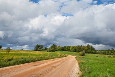 Gravel road, dirt track through Estonian countryside in Summer. Green fields and forest under dramatic sky with clouds.