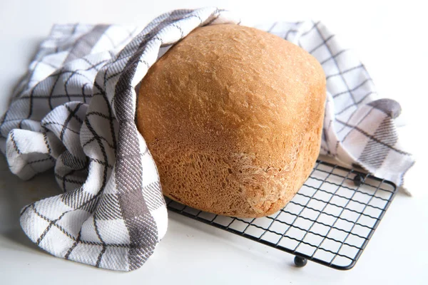 Home baked farmhouse mixed bread. Close-up on loaf of bread baked in baking machine. Whole wheat bread. Rustic organic farmhouse bio product on drying rack with stripy towel on white table.