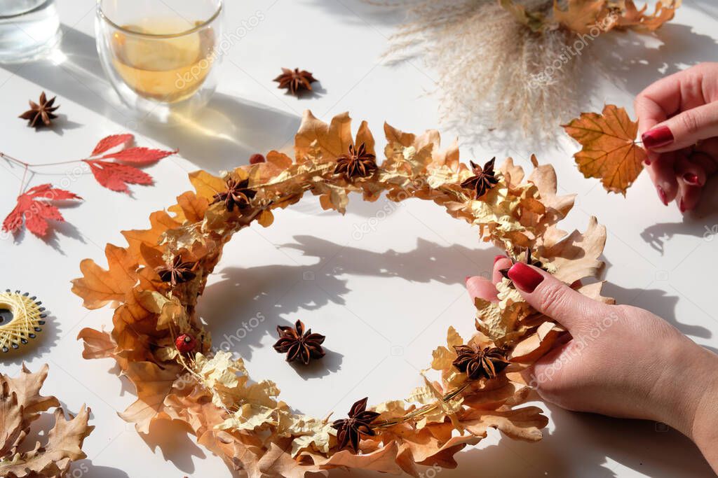 Hands making dried floral wreath from dry Autumn leaves and Fall berries. Hands with manicured nails fixing decorations with glue gun.