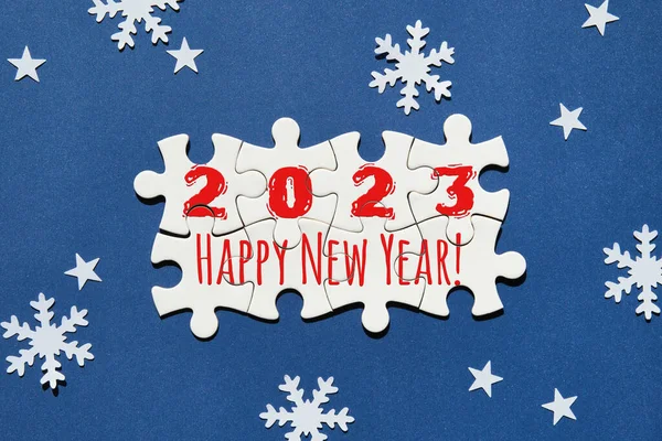 Jigsaw puzzle with numbers 2023 and caption text Happy New Year. Flat lay, top view on dark blue paper with snowflakes and star silhouettes. Simple minimal New Year greeting card design.