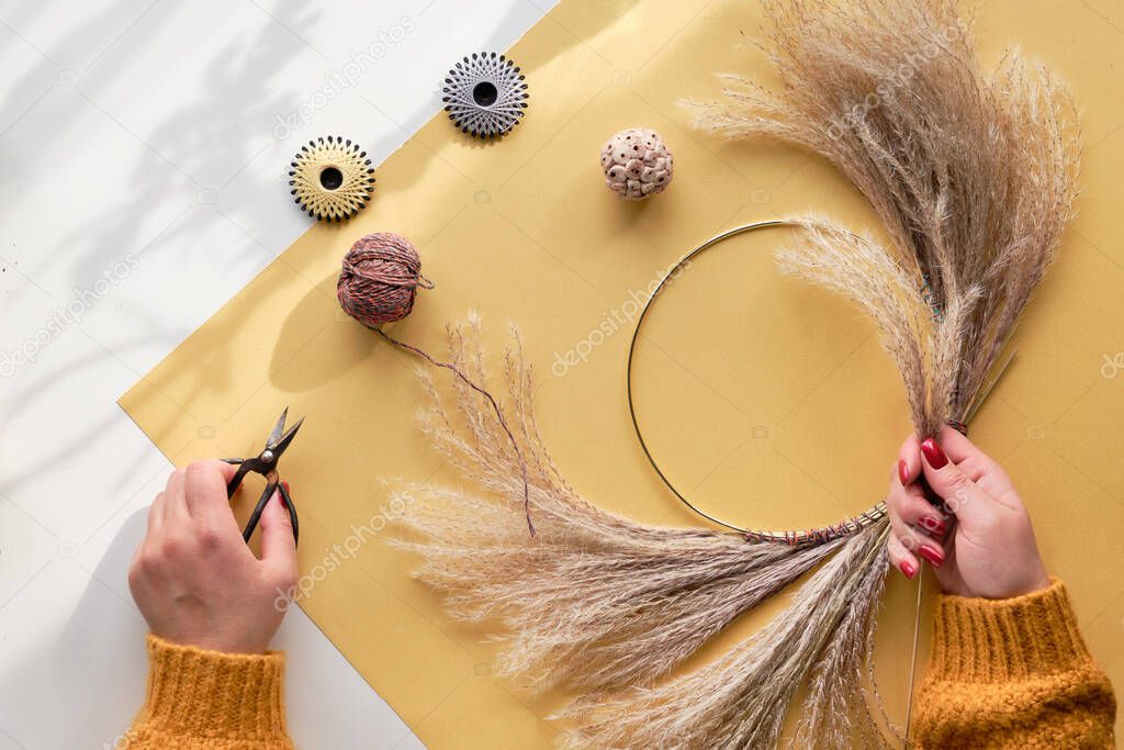 Hands making dried floral wreath from dry pampas grass and Autumn leaves. Hands in sweater tie decorations to metal frame. Flat lay on white yellow table, sunlight with long shadows.