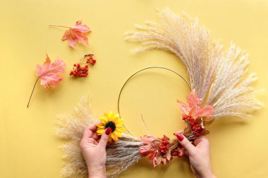 Kuru otlardan ve sonbahar yapraklarından kurumuş çiçek çelengi yapan eller. Süveter giymiş eller metal çerçeveye bağlanıyor. Düz sarı masa üzerinde yatıyordu, uzun gölgeli güneş ışığı..