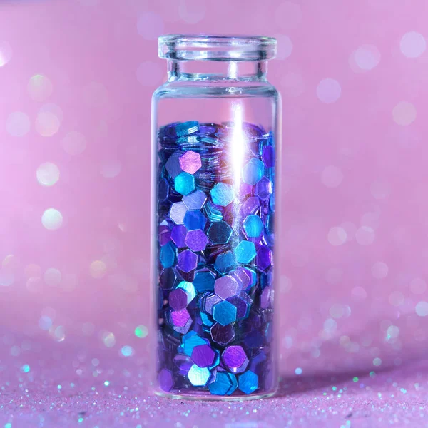 Glitter product in transparent bottle on pink sparkling background. Hexagone glitter in neon blue, purple and turquoise shades. Macro close-up on the bottle.
