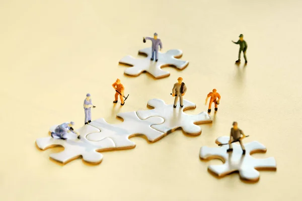 Team of tiny worker miniature figures on linked jigsaw puzzle pieces on golden yellow paper. Closeup on white jigsaw puzzle elements. Creative Team work in construction, development, logistics.