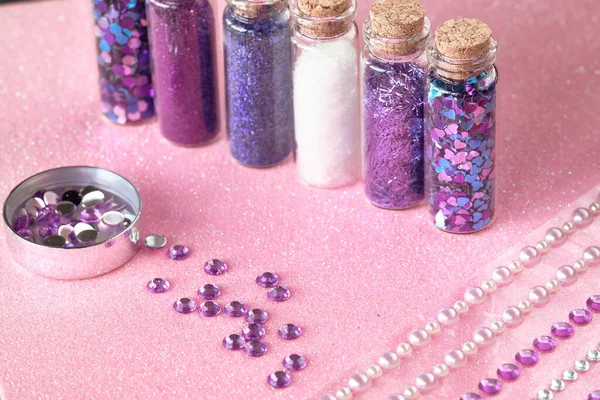 All kinds of glitter products on pink sparkling background. Close-up on vials, bottles with various glitter makeup in neon pink, blue and turquoise shades. Scattered strasses, rhinestones.