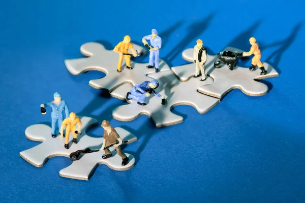 Team of tiny worker miniature figures on linked jigsaw puzzle pieces island on blue paper. Close-up on white jigsaw puzzle elements linked together and separate. Dramatic light with long shadows.